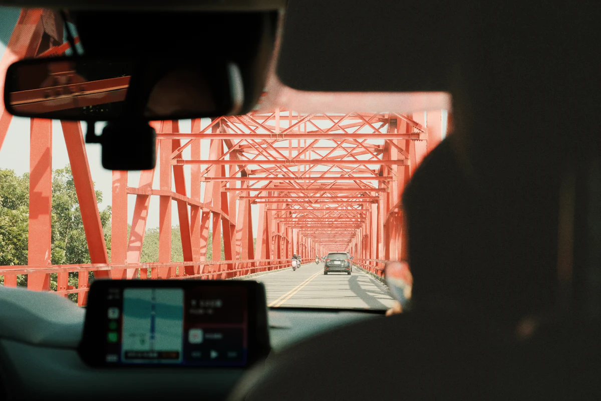 The view from the driver’s seat crossing a red steel bridge, with a navigation screen on the dash.