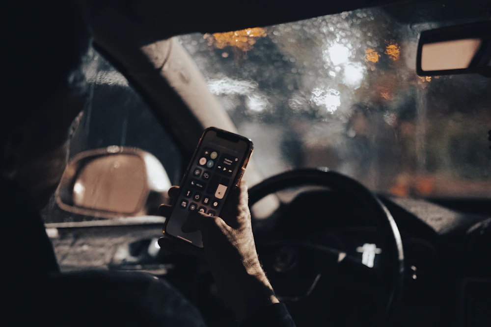 A driver sitting behind the wheel, holding a phone, on a rainy evening.