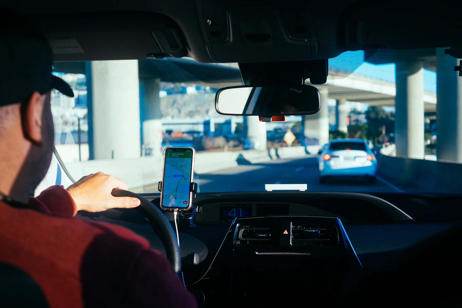 A driver in a car with a phone showing a navigation app mounted on the dashboard.